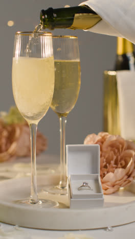 Vertical-Video-Of-Table-Set-For-Romantic-Marriage-Proposal-With-Champagne-Being-Poured-And-Engagement-Ring-In-Box